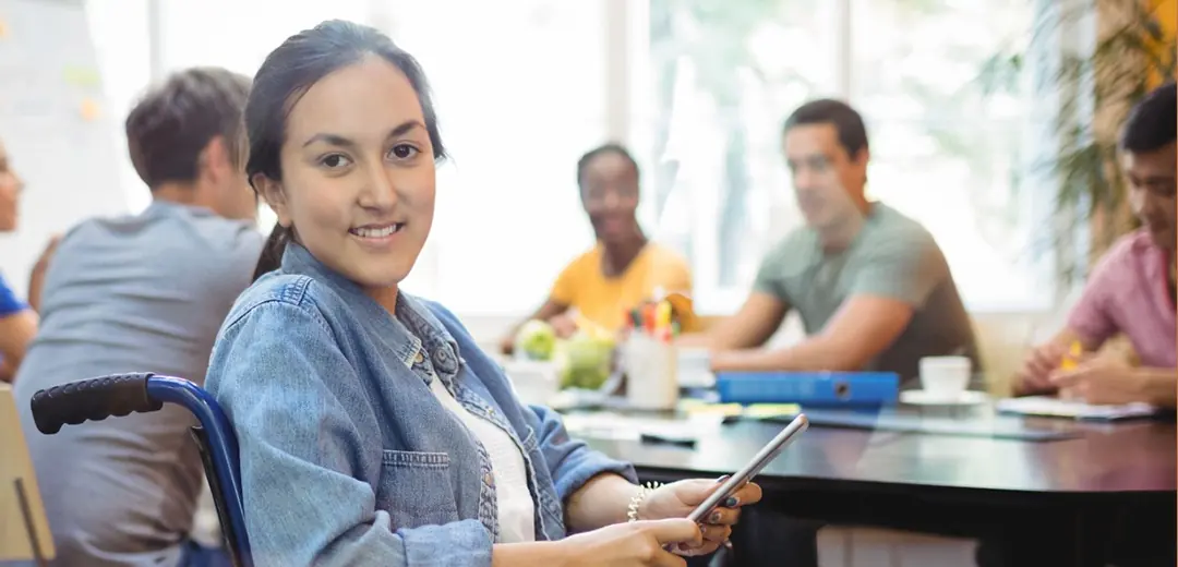 Joven en silla de ruedas sonriendo mientras usa una tableta en una reunión de trabajo.